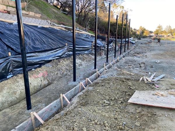 black tarp on the left over the hillside, footings to the left of a line of black fence posts that stretch into the back right, with the beginnings of